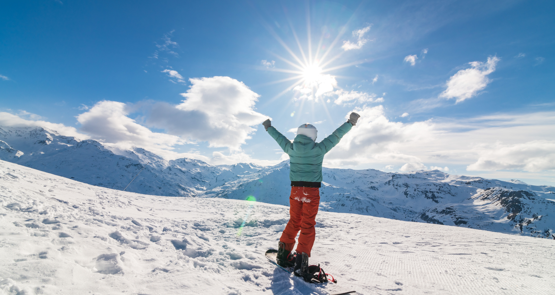 Un enfant de dos sur son snowboard levant les bras face aux montagnes enneigées