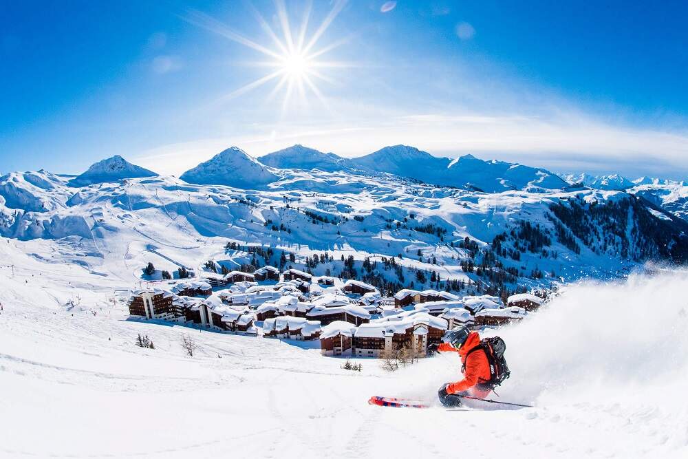 Un skieur dévalant les pistes de ski avec une station et les montagnes en arrière-plan
